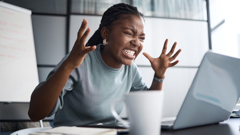 A frustrated person sitting at a desk looking at a laptop with a hand on their forehead.