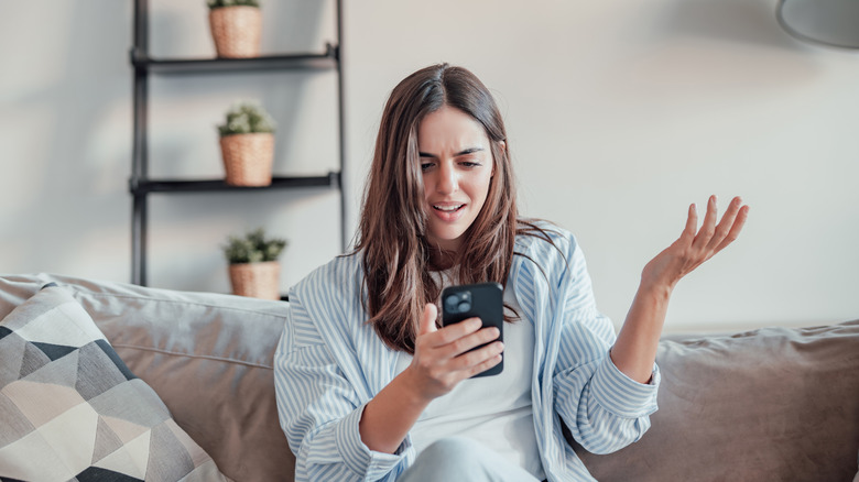 A woman sitting on a sofa looking at her smartphone with a look of extreme disappointment and frustration.