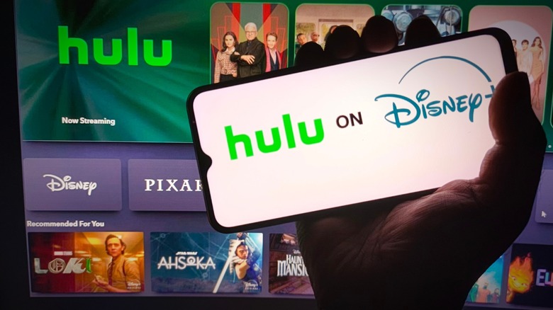 A phone displaying the Hulu app with the same interface reflected on a large television in the background.