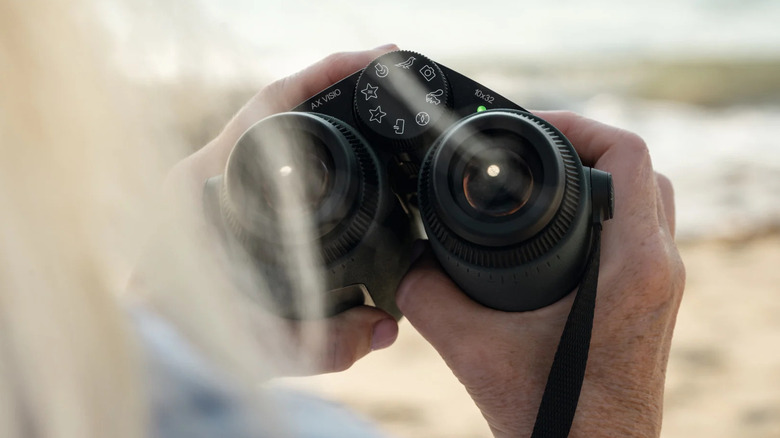 Close-up of hands holding the Swarovski AX Visio smart binoculars in an outdoor setting.