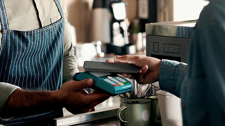 A person holding a Samsung phone near a contactless payment terminal in a coffee shop.