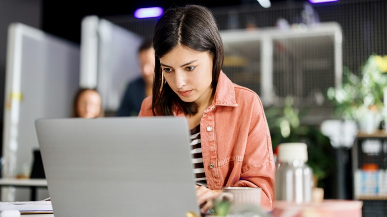 A woman in a pink jacket working on her laptop at a desk.