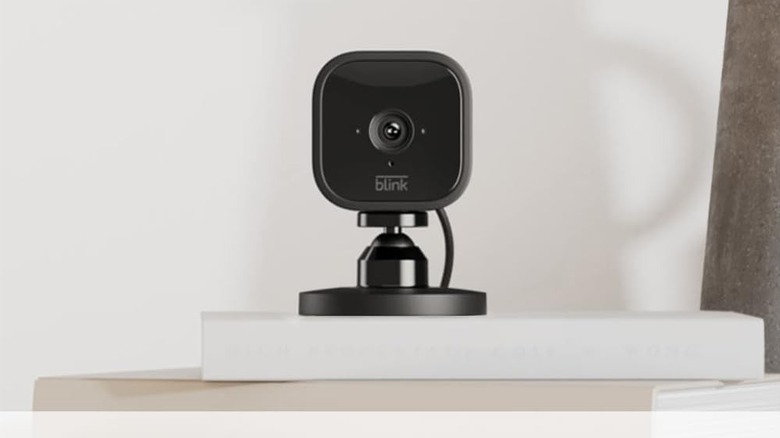 A black Blink security camera sitting on a white shelf in a living room.