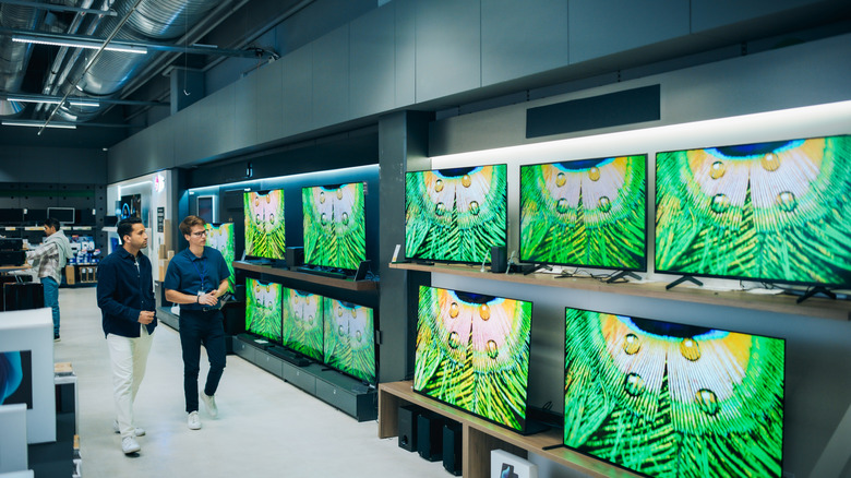Customers walking through a well-lit electronics store filled with various television displays.