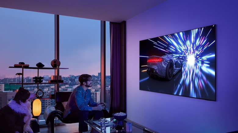 Two people sitting on a couch playing a high-speed racing game on a large OLED TV.