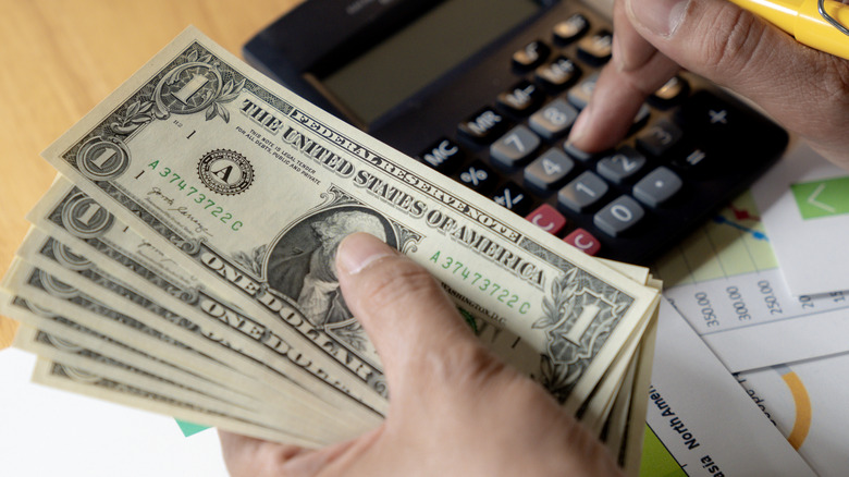 A close-up of a person using a calculator and counting US dollar bills near financial charts.
