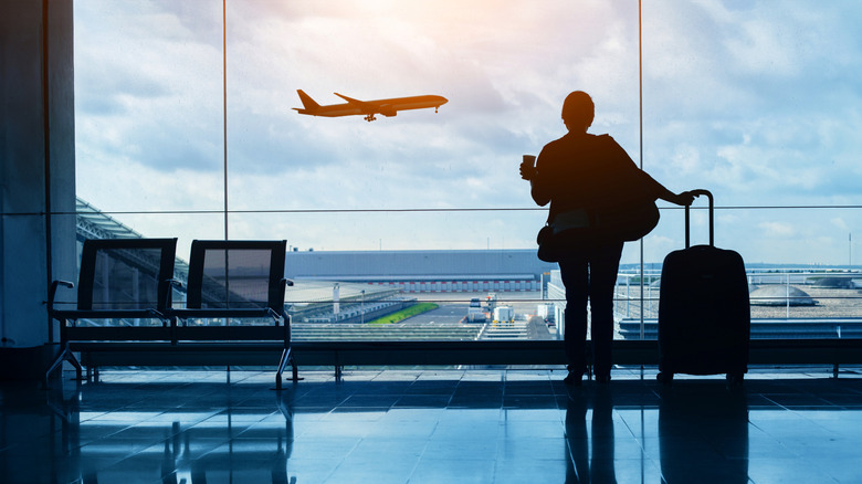 A silhouette of a passenger looking out at the tarmac from an airport terminal window.