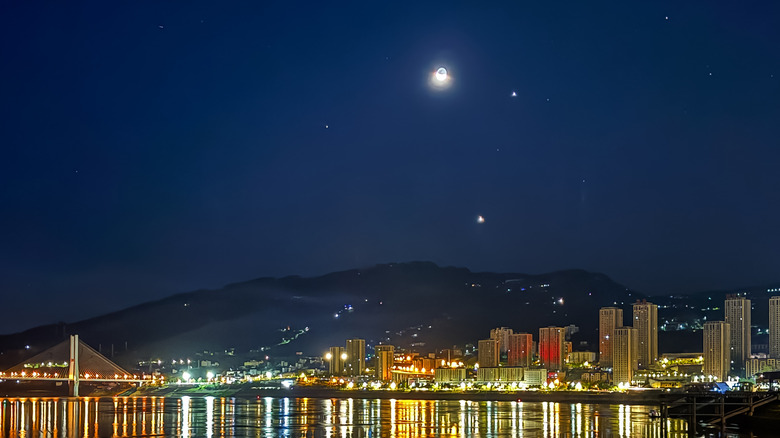 The crescent moon and bright planets Jupiter and Venus shining over a city skyline during twilight.