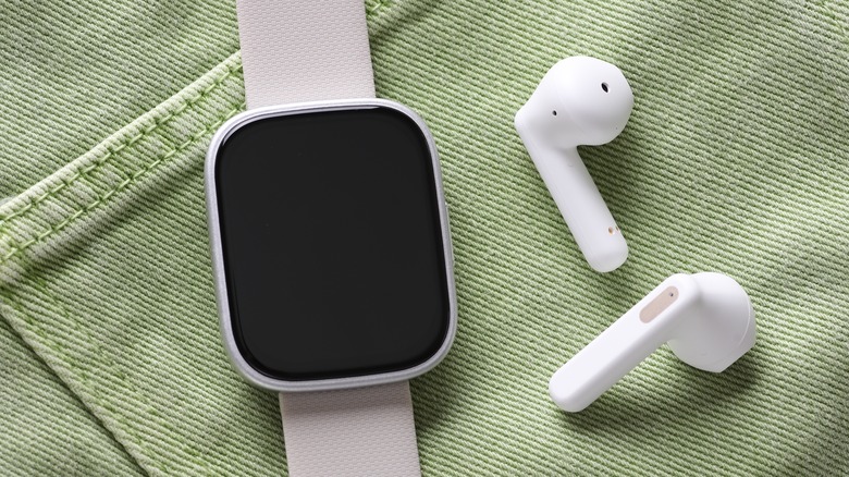A black smartwatch and wireless earbuds resting on green denim fabric.
