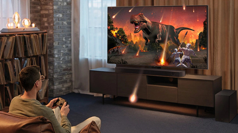 A person sitting on a couch gaming on a large TV equipped with a Samsung HW-Q600C soundbar and dedicated subwoofer.