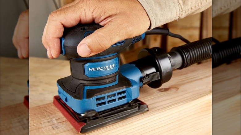 A worker using a Hercules 2.4 Amp orbital sheet sander on a piece of light-colored wood.