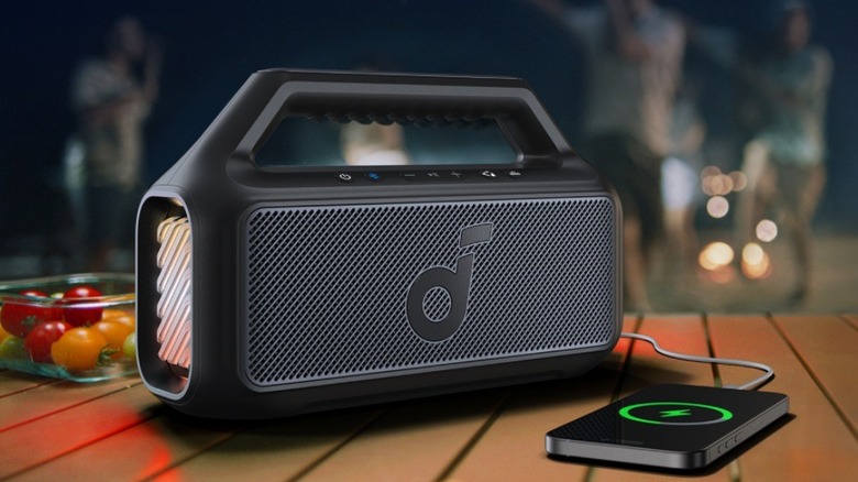 A Soundcore Boom V2 speaker sitting on a wooden surface while actively charging a mobile phone, highlighting its dual-purpose design. [2]
